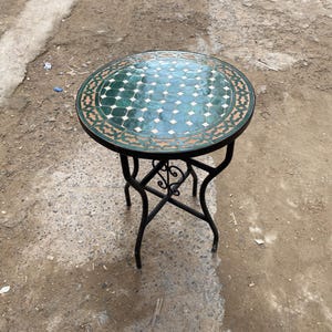 Może przedstawiać: Okrągły stół zewnętrzny z blatem z mozaiki w odcieniach zieleni i bieli. Stół ma czarną podstawę z kutego żelaza z dekoracyjnymi zawijasami. Stół stoi na betonowej powierzchni.