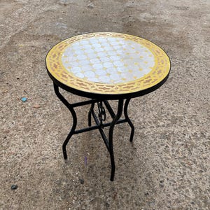 Może przedstawiać: Okrągły stół z mozaiki z czarną metalową ramą. Blat ma biało-żółty geometryczny wzór z żółtą obwódką. Stół ma trójnożną podstawę i nadaje się do użytku na zewnątrz.