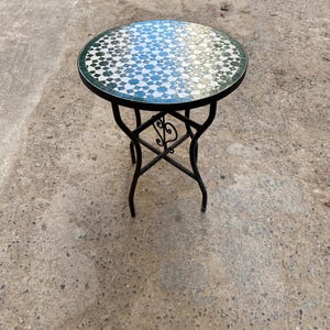 Może przedstawiać: Okrągły stolik boczny z blatem z mozaiki z geometrycznym wzorem w odcieniach niebieskiego, białego i zielonego. Stół ma czarną metalową ramę z dekoracyjnymi zawijasami i ma około 61 cm wysokości.