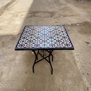 Può includere: Tavolo quadrato in mosaico con base in ferro battuto nero. Il piano presenta un colorato motivo geometrico nei toni del blu, verde, rosso e bianco. Il tavolo misura circa 76 cm quadrati.