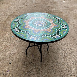 Może przedstawiać: Okrągły stół z mozaiki z czarną metalową ramą. Blat ma kolorowy geometryczny wzór w odcieniach niebieskiego, zielonego i żółtego. Stół ma średnicę około 61 cm.