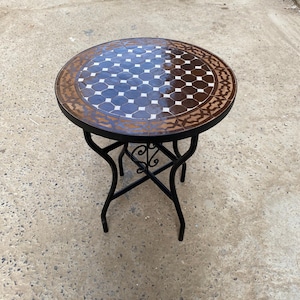 Może przedstawiać: Okrągły stół z mozaiki z ciemnobrązowym i białym wzorem na blacie. Stół ma czarną podstawę z kutego żelaza z dekoracyjnymi zawijasami. Stół ma średnicę około 61 cm.