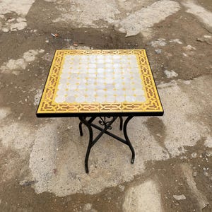 Może przedstawiać: Kwadratowy stół ogrodowy z blatem z mozaiki. Blat ma geometryczny wzór w kolorze białym, żółtym i brązowym. Stół ma czarną metalową ramę z dekoracyjnymi nogami. Stół ma wymiary około 61 cm x 61 cm.