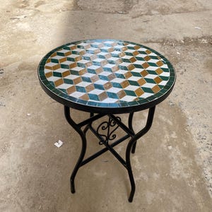 Może przedstawiać: Okrągły stół z mozaiką z czarną podstawą z kutego żelaza. Blat ma geometryczny wzór z zielonych, białych i złotych płytek. Stół ma średnicę około 61 cm i nadaje się do użytku na zewnątrz.