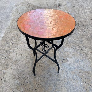 Może przedstawiać: Mały, okrągły stolik boczny z blatem z mozaiki w odcieniach pomarańczu i różu. Stół ma czarną metalową ramę z dekoracyjnymi zawijasami i zakrzywionymi nogami. Stół ma średnicę około 50 cm.