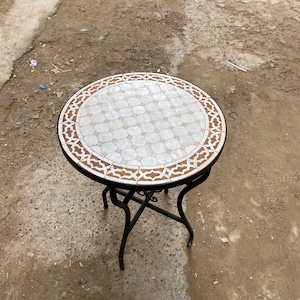 Może przedstawiać: Okrągły stół ogrodowy z blatem z mozaiki. Blat ma biało-brązowy geometryczny wzór. Stół ma czarną podstawę z kutego żelaza z zakrzywionymi nogami. Stół ma średnicę około 61 cm.
