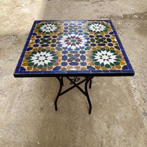 May include: Square mosaic tile table with a black metal frame. The tabletop features a colorful geometric pattern in shades of blue, green, white, and brown. The table is approximately 30 inches square.