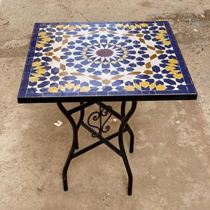 Może przedstawiać: Kwadratowy stół z mozaiki z czarną podstawą z kutego żelaza. Blat ma geometryczny wzór z niebieskich, żółtych i białych płytek, z ciemnobrązowym środkiem. Stół ma wymiary około 61 cm kwadratowych.