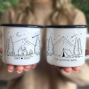 May include: Two white enamel camping mugs with black trim. One mug features a couple sitting by a campfire in front of a mountain with the text "James ♥ Ashley". The other mug features a tent in front of a mountain with the text "The Adventure Awaits".
