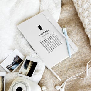 Könnte beinhalten: Ein weißes Notizbuch mit dem Wort "INTENTION" auf dem Einband und einem schwarzen Rechteck. Ein hellblauer Stift liegt auf dem Notizbuch, und mehrere kleine Fotos und eine Kamera befinden sich in der Nähe. Das Notizbuch hat ein graues Gummiband.