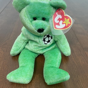 May include: A green plush bear toy with a soccer ball emblem on its chest. The bear has a red and white tag attached. The bear is sitting on a wooden surface.