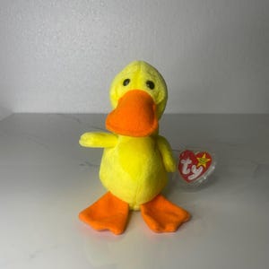 May include: A plush yellow duck toy with an orange beak and feet. The duck has black eyes and a red heart-shaped tag attached to its side. The toy is made of soft material and is designed to resemble a cartoon duck.