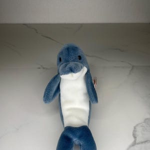 May include: A plush dolphin toy in shades of blue and white. The dolphin has a smiling face, flippers, and a tail. The toy is made of soft, fuzzy material and is designed to stand upright.