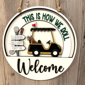 May include: Round wooden golf-themed welcome sign with a rope hanger. The sign features a golf cart, directional signs, and the phrases "This is how we roll" and "Welcome" in black and green lettering.
