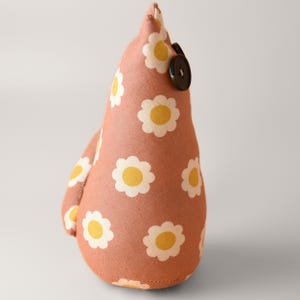 May include: A handmade fabric doorstop in a teardrop shape. The doorstop is covered in a peach-colored fabric with a repeating pattern of white daisies with yellow centers. A black button is sewn on the side.