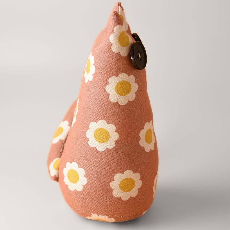 May include: A handmade fabric doorstop in a teardrop shape. The doorstop is covered in a peach-colored fabric with a repeating pattern of white daisies with yellow centers. A black button is sewn on the side.