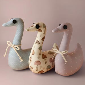 Puede incluir: Tres cisnes de tela rellenos. El cisne de la izquierda es azul claro con lunares blancos y una cinta color crema. El cisne central tiene un fondo crema con un estampado de setas marrones. El cisne de la derecha es rosa empolvado con lunares blancos y una cinta color crema.