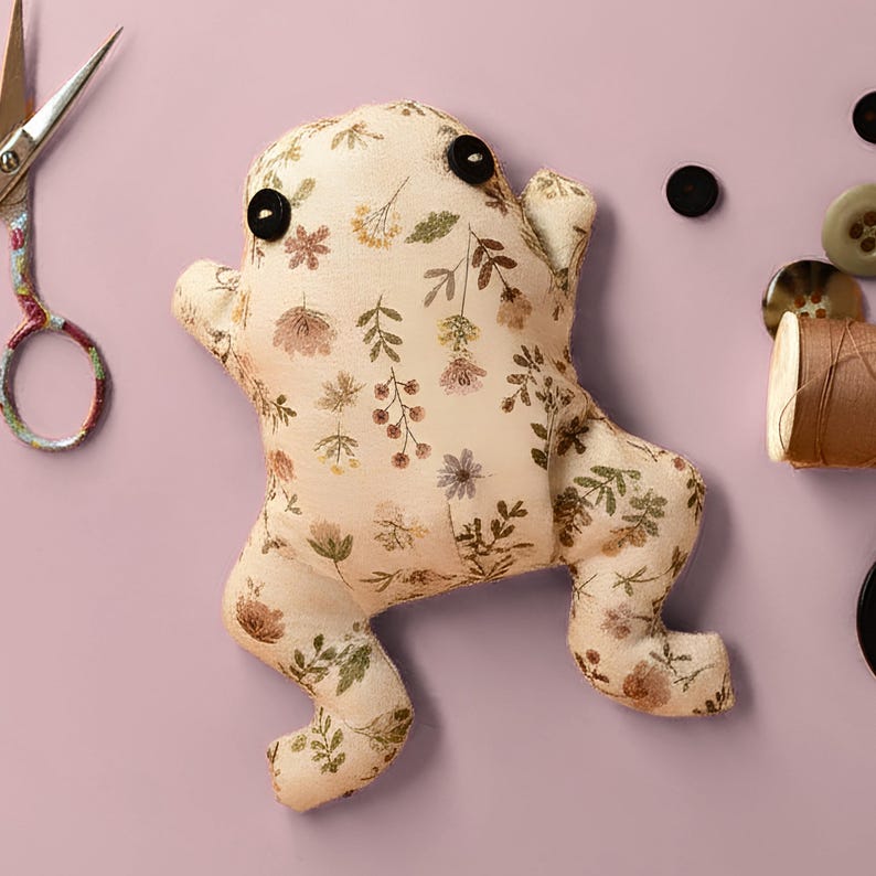 May include: A handmade frog-shaped plush toy with a floral pattern in cream, green, and pink. The frog has black button eyes and is surrounded by sewing notions, including scissors and buttons. The background is a solid lavender color.