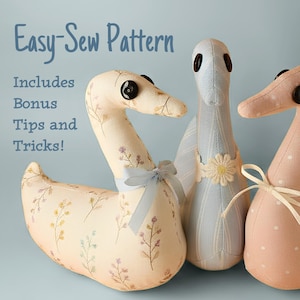 May include: Three fabric swan-shaped plush toys with button eyes. The left swan has a cream floral print and a light blue ribbon. The center swan is light blue with stripes and a flower applique. The right swan is solid pink with white dots.