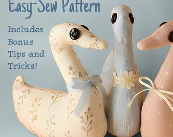 Può includere: Tre peluche in tessuto a forma di cigno con occhi a bottone. Il cigno a sinistra ha una stampa floreale color crema e un nastro azzurro. Il cigno centrale è azzurro a righe con un'applicazione floreale. Il cigno a destra è rosa tinta unita con pois bianchi.