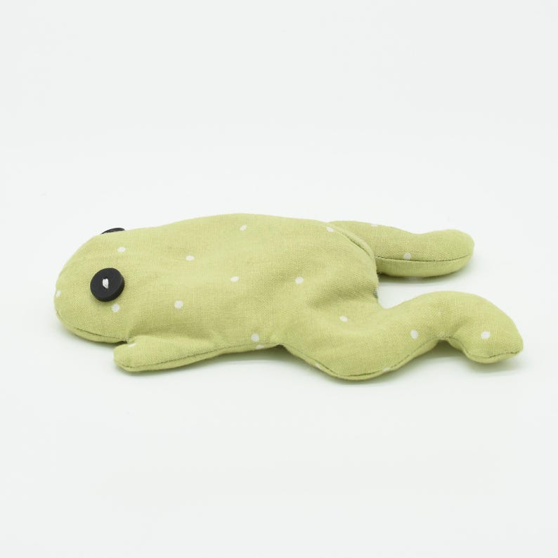 May include: A light green stuffed frog toy with white polka dots. The frog has two large black button eyes and is lying flat. The toy is made of soft fabric and is designed to look like a frog.