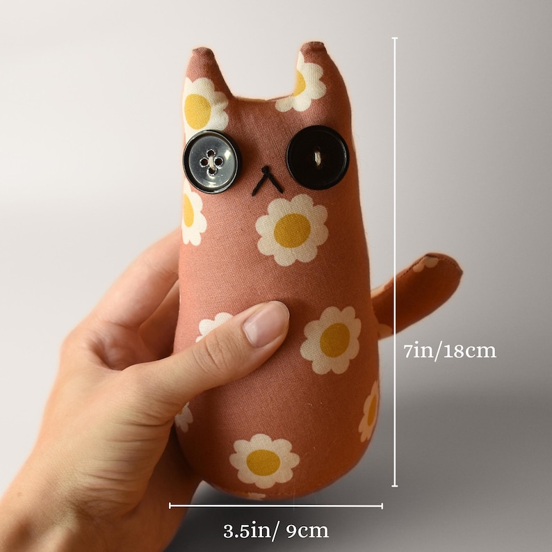 May include: A handmade plush cat doll, approximately 7 inches (18 cm) tall and 3.5 inches (9 cm) wide. The doll is made of a peach-colored fabric with a daisy pattern. It has button eyes and a stitched mouth.