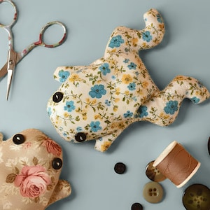 May include: Two handmade fabric whale-shaped toys with button eyes and floral patterns. One whale features blue and yellow flowers on a cream background, the other has pink roses. Scissors, buttons, and thread are scattered around.