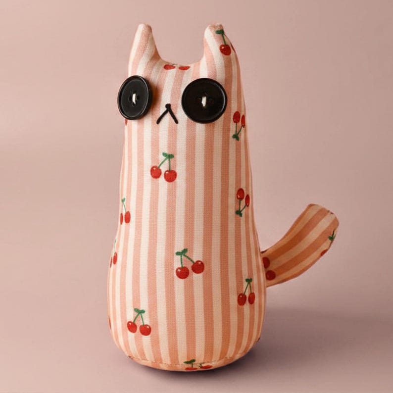 May include: A handmade cat-shaped plush toy with a pink and white striped pattern and red cherry accents. The cat has large black button eyes and a small black embroidered nose. The tail is curved. A whimsical and decorative item.