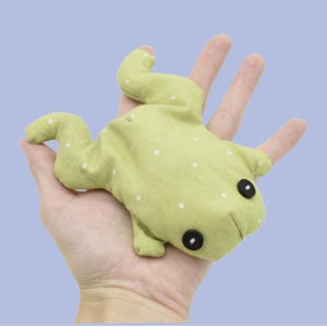 May include: A light green frog-shaped plush toy with white polka dots and black button eyes. The soft toy is made of fabric and has a stitched mouth. The frog is resting on a hand against a light blue background.