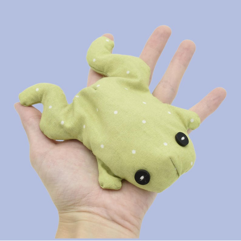 May include: A light green frog-shaped plush toy with white polka dots and black button eyes. The soft toy is made of fabric and has a stitched mouth. The frog is resting on a hand against a light blue background.
