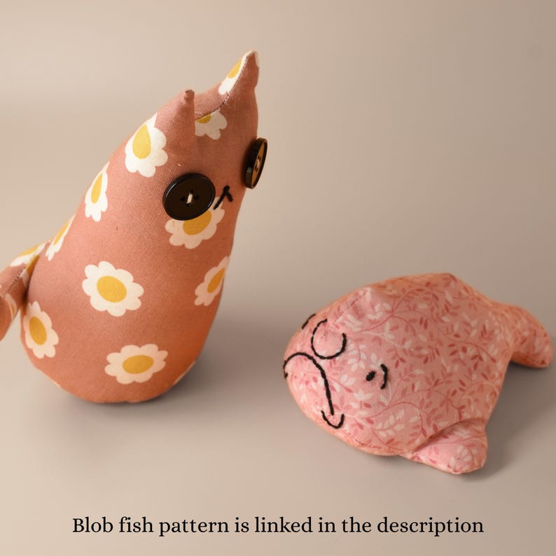 May include: Two handmade fabric toys. One is a pink blob-shaped fish with a whimsical face, and the other is a bird-like shape with a daisy pattern. The bird-like toy has button eyes. The text "Blob fish pattern is linked in the description" is at the bottom.
