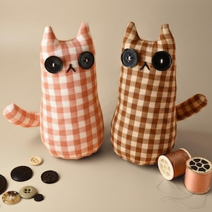 May include: Two handmade cat plushies with button eyes and stitched mouths. One cat is orange and white gingham, the other is brown and white gingham. Several buttons and spools of thread are scattered around the cats, suggesting a craft or sewing theme.