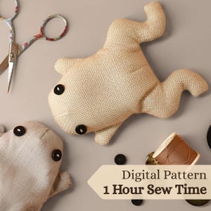 May include: Two beige fabric ghost-shaped plush toys with black button eyes. The image also shows a pair of scissors, black buttons, and a spool of brown thread. Text on the image reads "Digital Pattern" and "1 Hour Sew Time".