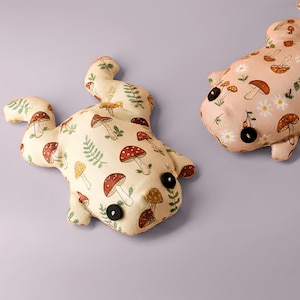 May include: Two whimsical frog-shaped pillows. One is cream-colored with a pattern of red-capped mushrooms and green ferns. The other is pink with a similar mushroom design and daisies. Both have black button eyes.