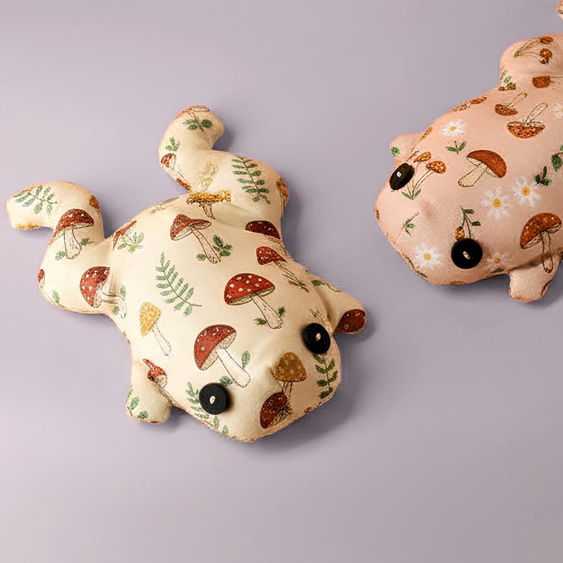 May include: Two whimsical frog-shaped pillows. One is cream-colored with a pattern of red-capped mushrooms and green ferns. The other is pink with a similar mushroom design and daisies. Both have black button eyes.