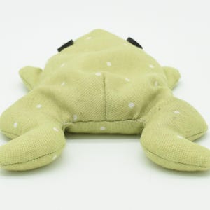 May include: A light green frog-shaped plush toy with white polka dots. The toy has black eyes and outstretched legs. The frog is made of soft fabric and is lying on a white surface.