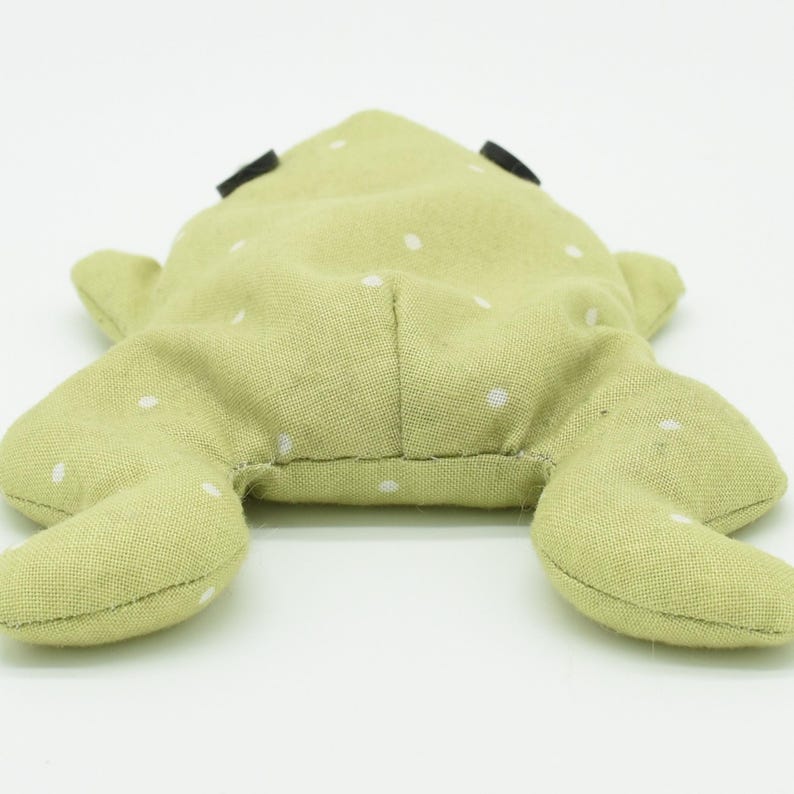 May include: A light green frog-shaped plush toy with white polka dots. The toy has black eyes and outstretched legs. The frog is made of soft fabric and is lying on a white surface.