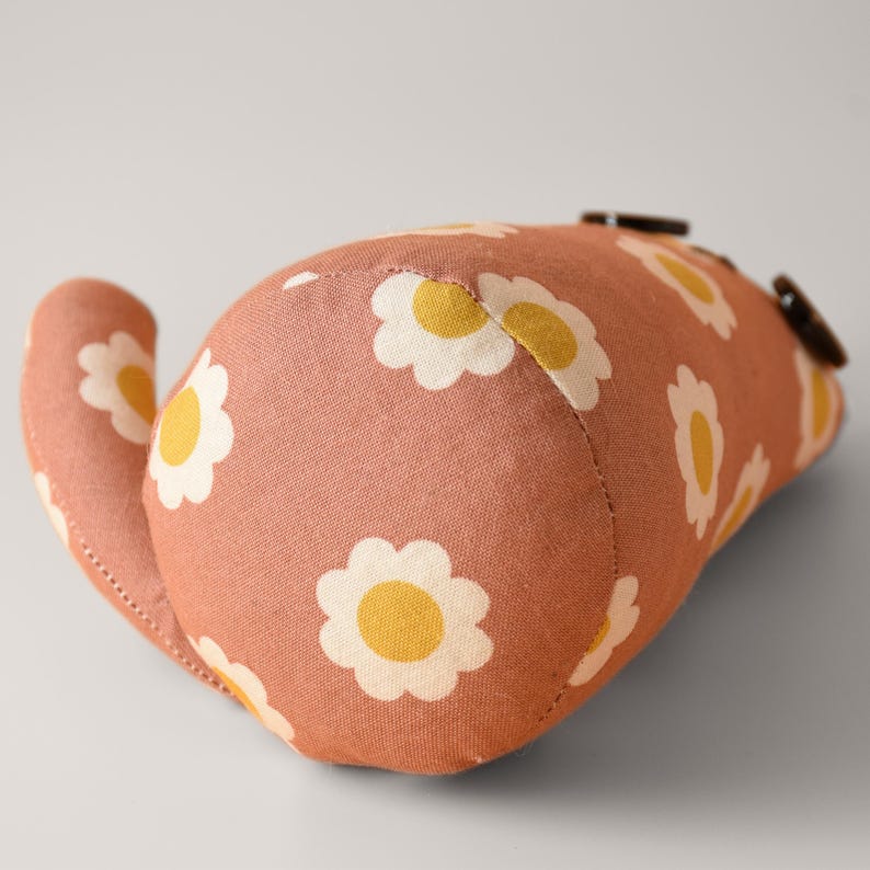 May include: A soft, peach-colored fabric cat toy with a daisy pattern in white and yellow. The toy has a curved shape with a tail and two brown button accents. The fabric appears to be cotton or a similar material.