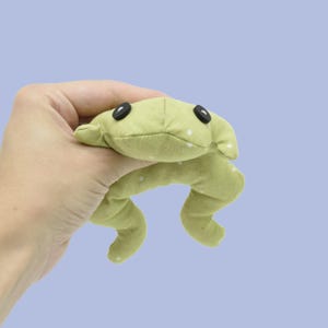 May include: A light green frog-shaped plush toy with white polka dots and black button eyes. The soft toy is made of fabric and has curved legs. The frog is held in a hand against a light blue background.