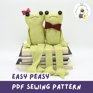 Peut inclure: Deux peluches en forme de grenouilles vertes à pois blancs, l'une avec un nœud papillon rouge et l'autre avec un nœud rouge. Elles sont assises sur une pile de livres. Le texte "EASY PEASY PDF SEWING PATTERN" est en bas.