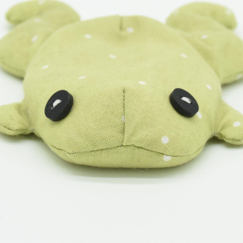 May include: A light green, plush frog-shaped toy with white polka dots. The frog has two black button eyes and small, rounded arms and legs. The toy is made of soft fabric.