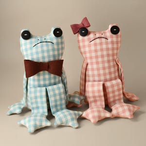 May include: Two stuffed frog toys made of gingham fabric. The left frog is light blue and white with a brown felt bowtie. The right frog is pink and white with a small pink bow on its head. Both have black button eyes and stitched mouths.