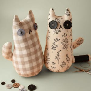 May include: Two handmade cat plush toys. One cat is tan and white checkered, the other is beige with a black floral pattern. Both have button eyes and stitched noses. Sewing notions, including buttons and scissors, are scattered around.