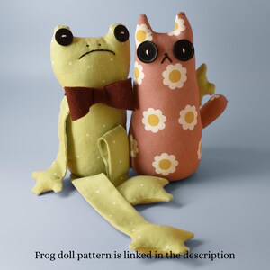 May include: Two handmade plush dolls: a green frog with a brown bow tie and a pink cat with daisy patterns. Both dolls have button eyes. The frog doll has long legs. Text at the bottom reads: "Frog doll pattern is linked in the description."