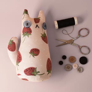 May include: A handmade cat-shaped plush toy with a strawberry pattern, blue button eyes, and a stitched mouth. Accessories include black thread, small scissors, and various buttons. The cat is approximately 8 inches tall.