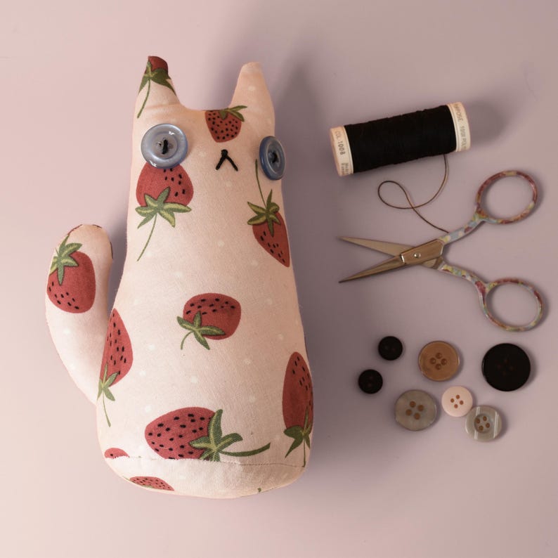 May include: A handmade cat-shaped plush toy with a strawberry pattern, blue button eyes, and a stitched mouth. Accessories include black thread, small scissors, and various buttons. The cat is approximately 8 inches tall.