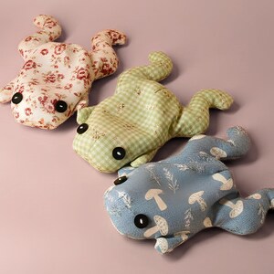 May include: Three handmade frog plush toys in a row. The top frog is floral, the middle is green gingham, and the bottom is blue with a mushroom pattern. Each frog has large black button eyes and is lying on a pink surface.