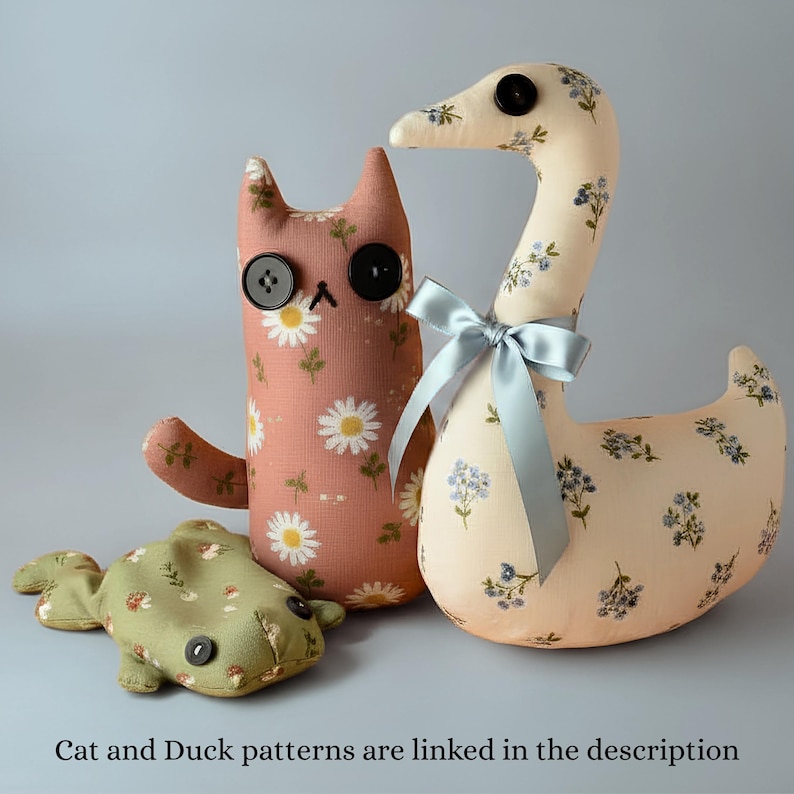 May include: Three handmade fabric toys: a cat, a duck, and a fish. The cat is pink with white daisy print and button eyes. The duck is cream-colored with blue floral print and a blue ribbon. The fish is green with a floral pattern. Text: "Cat and Duck patterns are linked in the description."