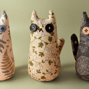 May include: Three handmade cat-shaped fabric dolls. The dolls have button eyes and stitched features. The doll on the left is beige with a fern pattern. The center doll is beige with a floral pattern. The doll on the right is dark gray with a floral pattern.