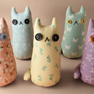 May include: Five plush cat-shaped toys made of patterned fabric. Each toy features two button eyes, a stitched nose, and pointed ears. The fabrics include light blue with white snowflakes, yellow with green floral prints, pink with white flowers, light green with white butterflies, and purple with white stars and moons.